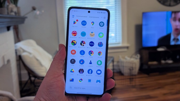 Рука, що містить смартфон Poco F7 Pro, що відображає домашній екран додатка з різними барвистими піктограмами додатків, з телевізором на задньому плані, що показує людину, яка говорить.
