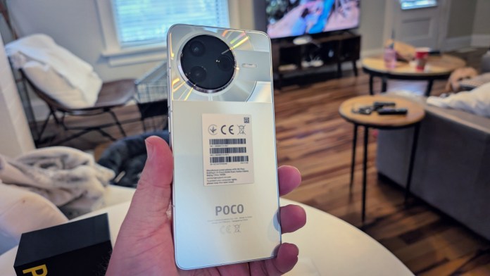 Людина, що тримає блискучу спинку смартфона Poco F7 Pro, розкриває його модуль камери та брендинг із затишною налаштуванням вітальні на задньому плані.